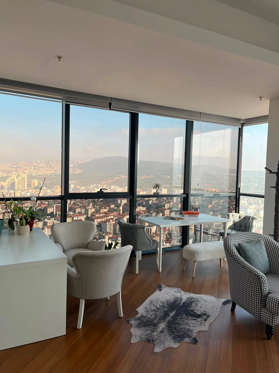 Köşe cam cephesinden şehir panoraması; beyaz masanın çevresinde açık gri koltuklar ve ahşap zeminde açık renkli halı.
